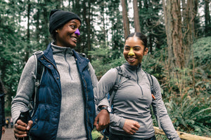 A smiling mother and daughter hiking along a forest trail and wearing purple and yellow shades of Nöz's eco friendly sunscreen. This family is in gray jackets and backpacks. Their arms are interlocked.